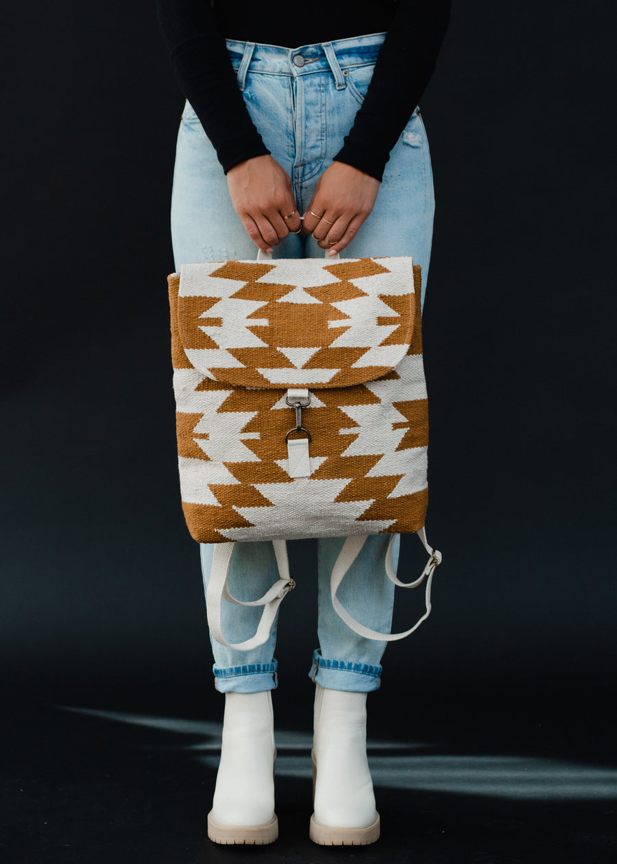 A person in light blue jeans, a black long-sleeve shirt, and white boots holds the panacheaccessories Inez Backpack, featuring white and brown geometric designs and adjustable straps, against a plain black background.