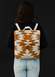 A person with wavy brown hair, in a black top and light blue jeans, is seen from behind carrying the Inez Backpack by panacheaccessories, featuring adjustable straps and a brown and white geometric pattern on black.