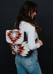 A woman in a white long-sleeve shirt and blue jeans smiles while wearing the Collins Backpack by panacheaccessories, which features adjustable straps and a red, white, and gray geometric Aztec pattern, against a black background.