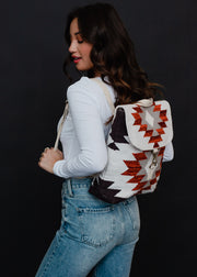 A woman with long dark hair in a white long-sleeve shirt and blue jeans poses sideways with the panacheaccessories Collins Backpack, featuring a red, white, and black Aztec geometric pattern and adjustable straps against a dark background.