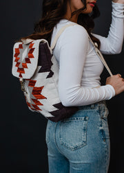 A woman in a white long-sleeve shirt and blue jeans holds the panacheaccessories Collins Backpack, featuring adjustable straps and a bold red, black, and white Aztec-inspired geometric design against a dark background.