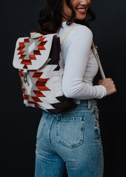 A woman in blue jeans and a white long-sleeve top smiles while modeling the Collins Backpack by panacheaccessories, which features adjustable straps and a red, white, and black Aztec geometric pattern against a black background.