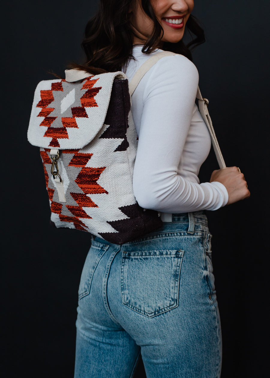 A woman in blue jeans and a white long-sleeve top smiles while modeling the Collins Backpack by panacheaccessories, which features adjustable straps and a red, white, and black Aztec geometric pattern against a black background.