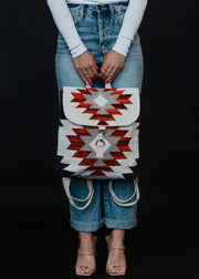 A person in a white long-sleeve shirt and blue jeans holds the Collins Backpack by panacheaccessories, featuring red, white, and black Aztec-inspired geometric designs and adjustable straps. They wear nude heeled sandals against a black background.