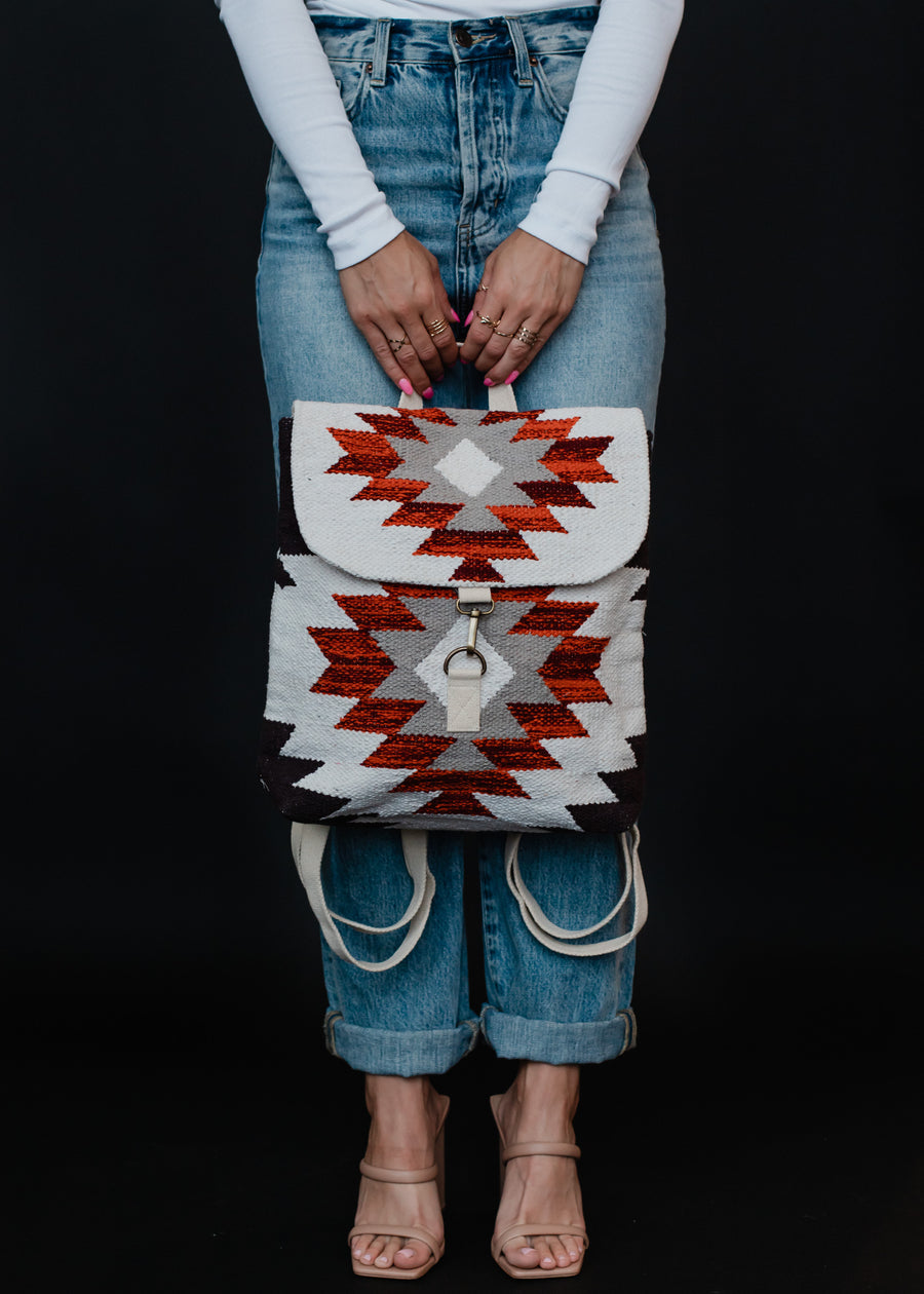 A person in a white long-sleeve shirt and blue jeans holds the Collins Backpack by panacheaccessories, featuring red, white, and black Aztec-inspired geometric designs and adjustable straps. They wear nude heeled sandals against a black background.