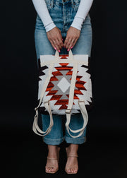 Wearing rolled-up blue jeans, a white long-sleeve top, and nude heeled sandals, a person holds the panacheaccessories Collins Backpack featuring adjustable straps and a bold red, black, and white geometric southwestern pattern on black.