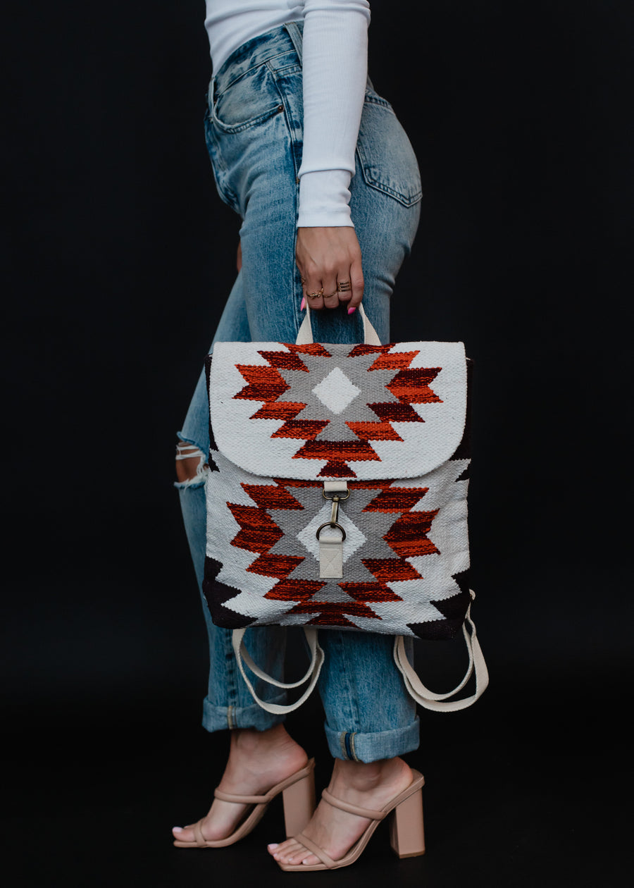 A person in a white long-sleeve top, ripped blue jeans, and nude heeled sandals holds the panacheaccessories Collins Backpack featuring adjustable straps and a red, white, and gray geometric Aztec pattern against a black background.