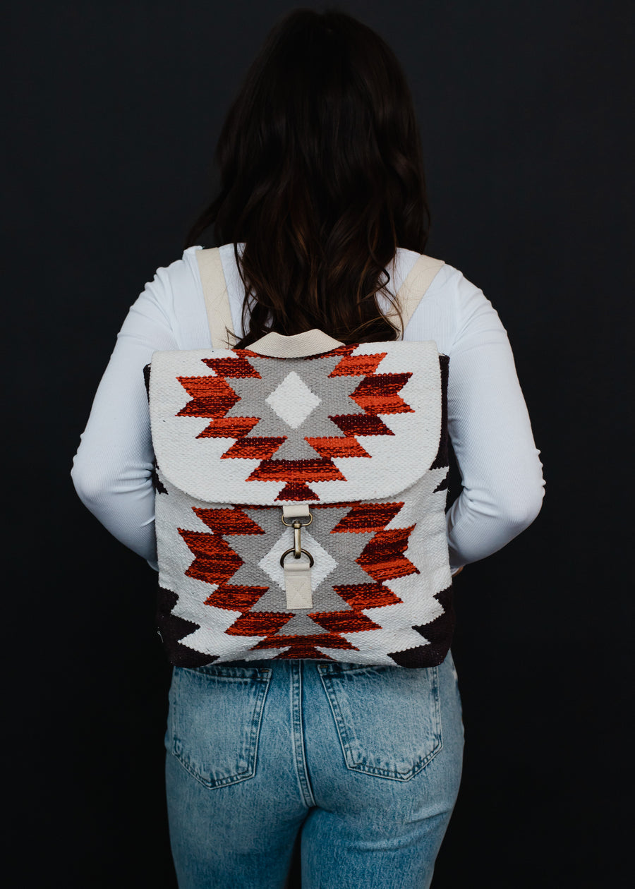 A person with long dark hair, in a white shirt and blue jeans, stands facing away, showcasing the panacheaccessories Collins Backpack featuring adjustable straps and a bold red, white, and gray geometric design on black.
