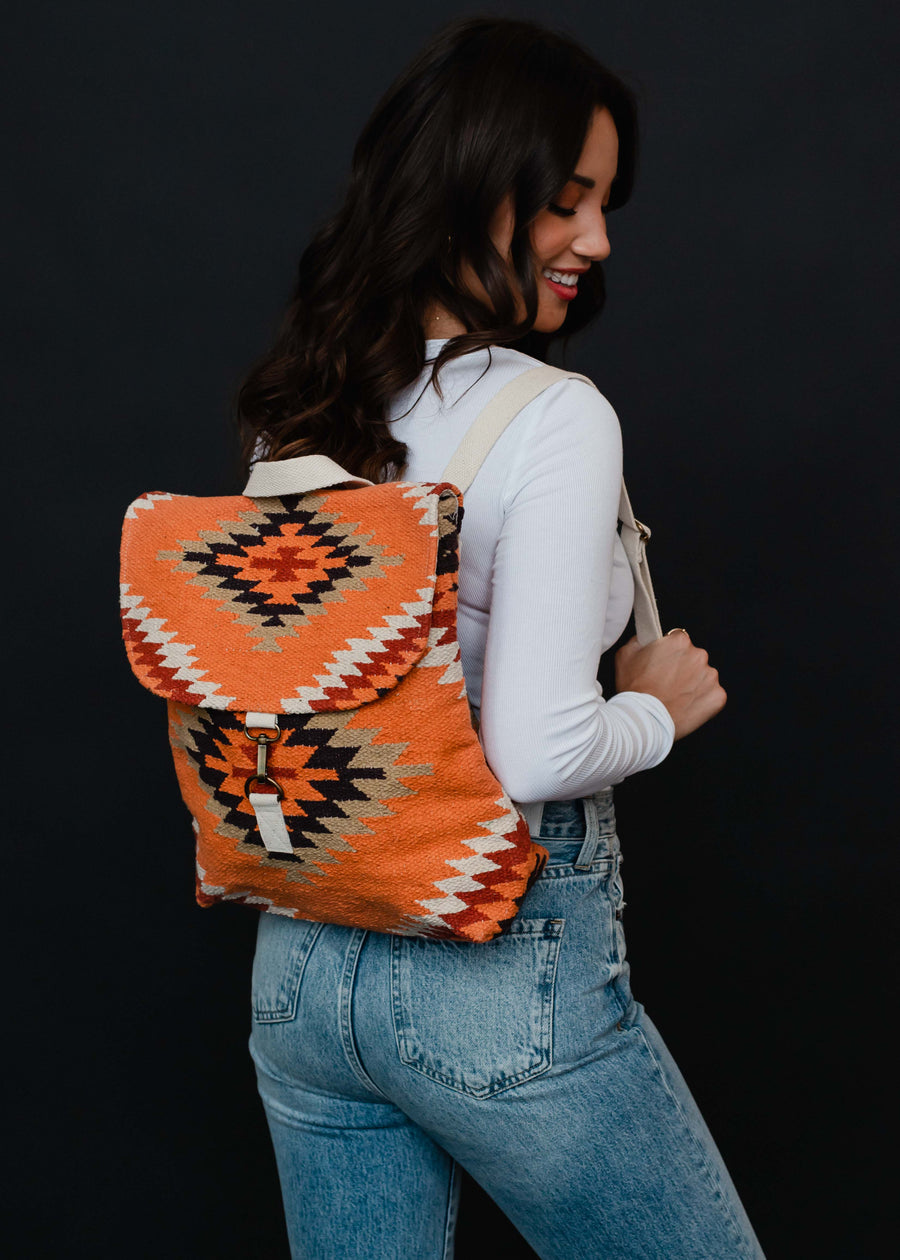 A woman with long dark hair, in a white long-sleeve top and light blue jeans, poses sideways against a black background with the panacheaccessories Savannah Backpack, which features an Aztec print and adjustable straps.