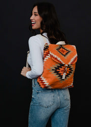A woman with long dark hair in a white long-sleeve shirt and blue jeans smiles while carrying the Savannah Backpack by panacheaccessories, featuring a geometric Aztec design and adjustable straps, set against a black background.