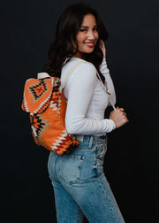 A smiling woman with long dark hair, in a white long-sleeve shirt and blue jeans, poses sideways with the Savannah Backpack by panacheaccessories, featuring adjustable straps, against a dark background.