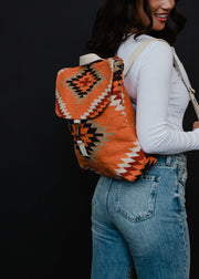 A woman in a white long-sleeve top and blue jeans smiles while carrying the panacheaccessories Savannah Backpack, featuring adjustable straps and a southwestern Aztec design, against a black background.