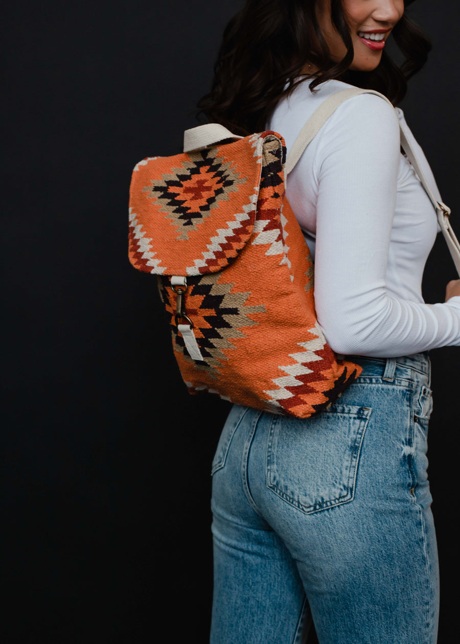 A woman in a white long-sleeve top and blue jeans smiles while carrying the panacheaccessories Savannah Backpack, featuring adjustable straps and a southwestern Aztec design, against a black background.