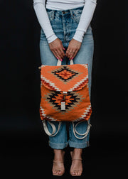 A person in a white long-sleeve shirt, blue jeans, and beige heeled sandals holds the orange Savannah Backpack by panacheaccessories, featuring geometric Aztec designs and adjustable straps, against a black background.
