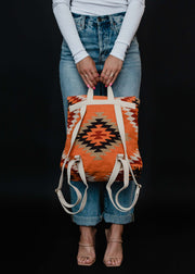 A person in a white long-sleeve top and blue jeans holds the Savannah Backpack by panacheaccessories, featuring orange, black, and cream Aztec-inspired geometric patterns with beige adjustable straps. The background is black.