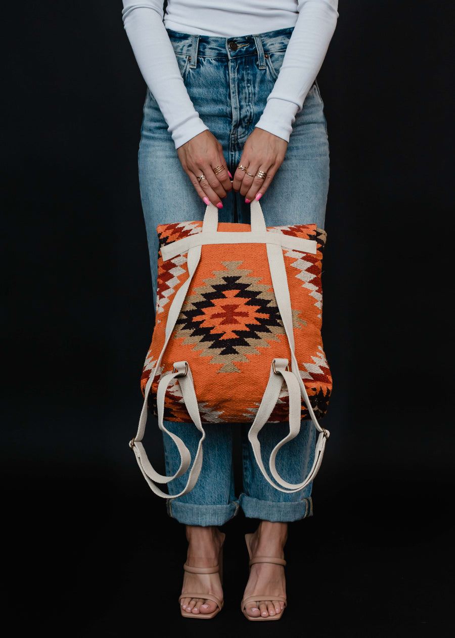 A person in a white long-sleeve top and blue jeans holds the Savannah Backpack by panacheaccessories, featuring orange, black, and cream Aztec-inspired geometric patterns with beige adjustable straps. The background is black.