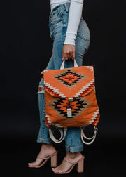A person in blue jeans, a white long-sleeve top, and nude heeled sandals holds the panacheaccessories Savannah Backpack featuring geometric black, red, and white designs against a black background.