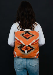 A person with long brown hair, dressed in a white top and blue jeans, is seen from behind carrying the Savannah Backpack by panacheaccessories, featuring adjustable straps and a geometric pattern on black.
