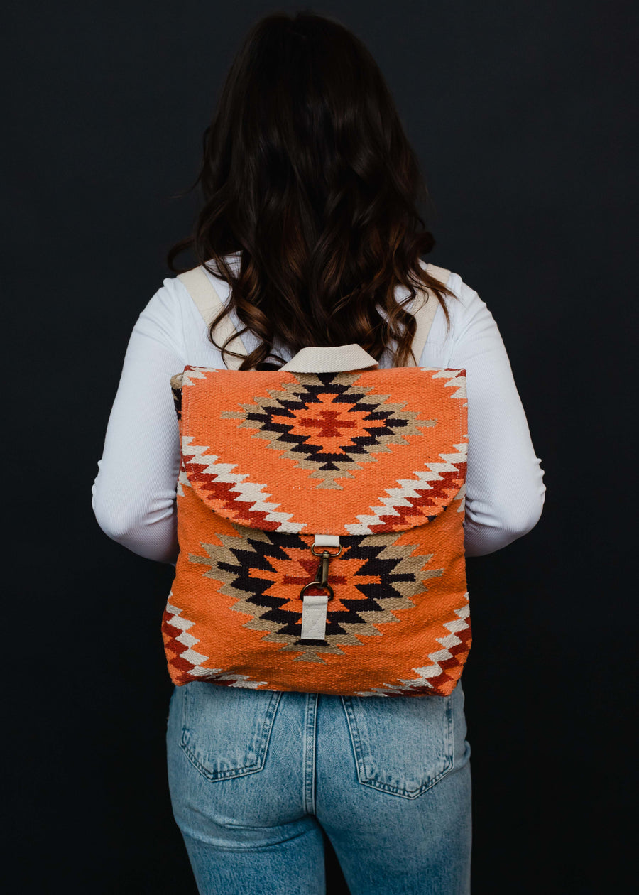 A person with long brown hair, dressed in a white top and blue jeans, is seen from behind carrying the Savannah Backpack by panacheaccessories, featuring adjustable straps and a geometric pattern on black.