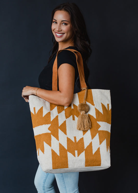A smiling woman in a black top and light jeans holds the Easy Street Tote by panacheaccessories, featuring tan and white geometric patterns, a camel pom tassel, and leather suede straps, against a dark background.