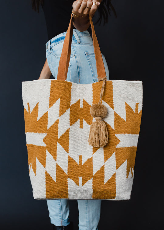 A person in light blue jeans holds the Easy Street Tote by panacheaccessories, featuring leather suede straps and a geometric white and ochre pattern with a camel pom tassel keychain, set against a solid black background.