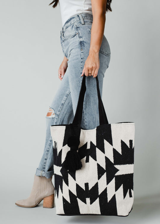 A woman in light-wash jeans, a white top, and beige boots holds the panacheaccessories Londyn Tote—featuring a bold black-and-white geometric pattern, leather suede straps, and a black pom tassel—against a plain gray background.