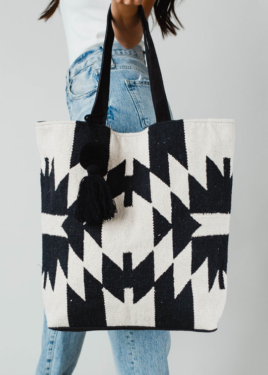 A person in light blue jeans and a white top holds the Londyn Tote by panacheaccessories, featuring black and white geometric patterns, suede leather straps, and black tassel accents, set against a plain background.