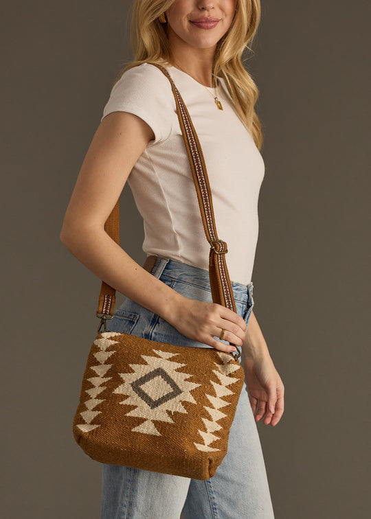 A woman in a white short-sleeve top and light blue jeans carries the panacheaccessories Tatum Crossbody, featuring a white geometric pattern and an adjustable strap.