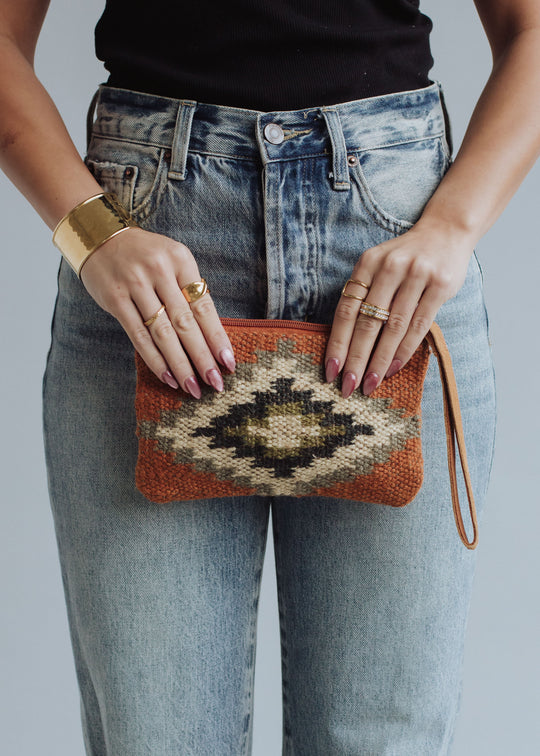 A person in blue jeans and a black top holds the panacheaccessories Veronica Wristlet, a small western-inspired clutch with an orange, cream, black, and gray geometric pattern and leather pull. They wear gold rings and a bracelet.