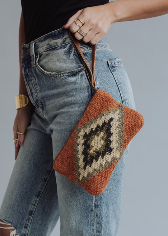 A person in high-waisted, faded blue jeans and a black top holds the panacheaccessories Veronica Wristlet, featuring a western-inspired geometric pattern in earthy tones with a leather pull, accessorized with gold rings and a bracelet.