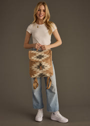 A smiling woman stands against a plain brown background holding the Zola Backpack by panacheaccessories, featuring adjustable straps, while wearing a white t-shirt, light blue jeans, and white sneakers.