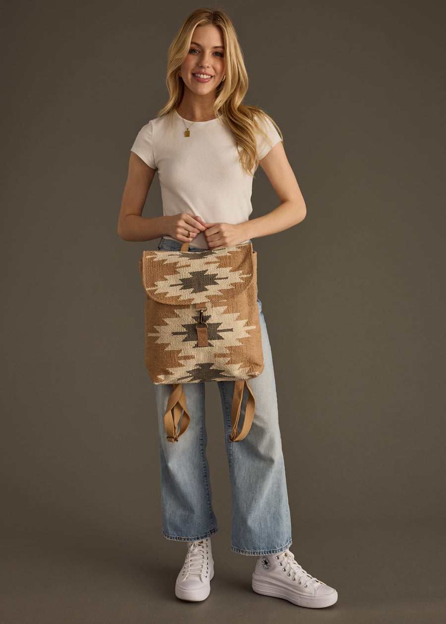 A smiling woman stands against a plain brown background holding the Zola Backpack by panacheaccessories, featuring adjustable straps, while wearing a white t-shirt, light blue jeans, and white sneakers.