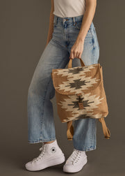 A person wearing light blue jeans, a white shirt, and white Converse holds the Zola Backpack by panacheaccessories—a brown cotton bag with geometric patterns and adjustable straps—against a plain background.