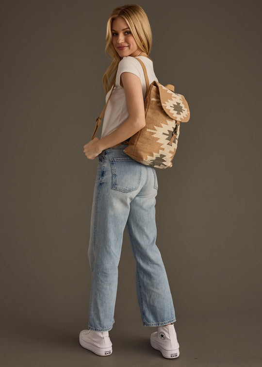 A woman with long blonde hair stands sideways against a plain background, smiling as she looks back over her shoulder. She wears a white T-shirt, light blue jeans, white sneakers, and carries the Zola Backpack by panacheaccessories.