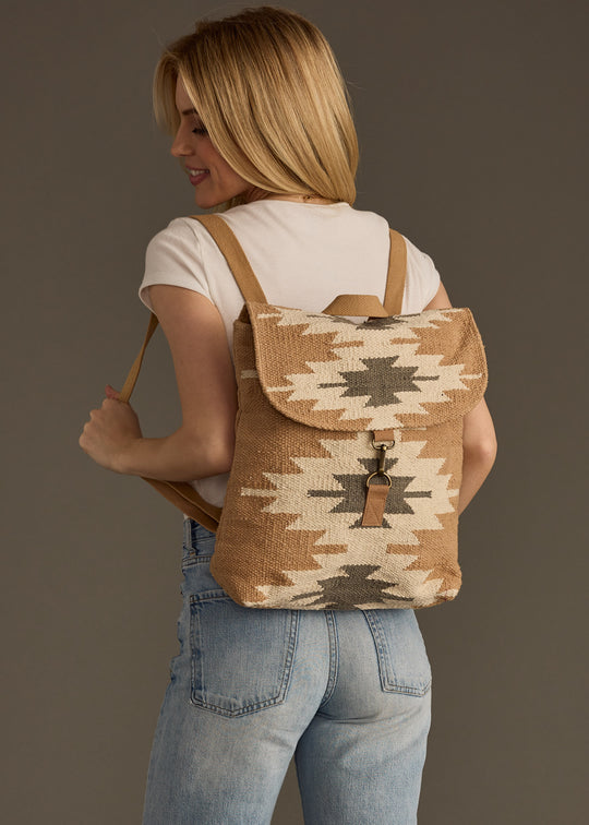 A woman in a white tee and light blue jeans smiles sideways with the Zola Backpack by panacheaccessories—featuring a tan and cream western pattern and adjustable straps—on her back against a plain background.