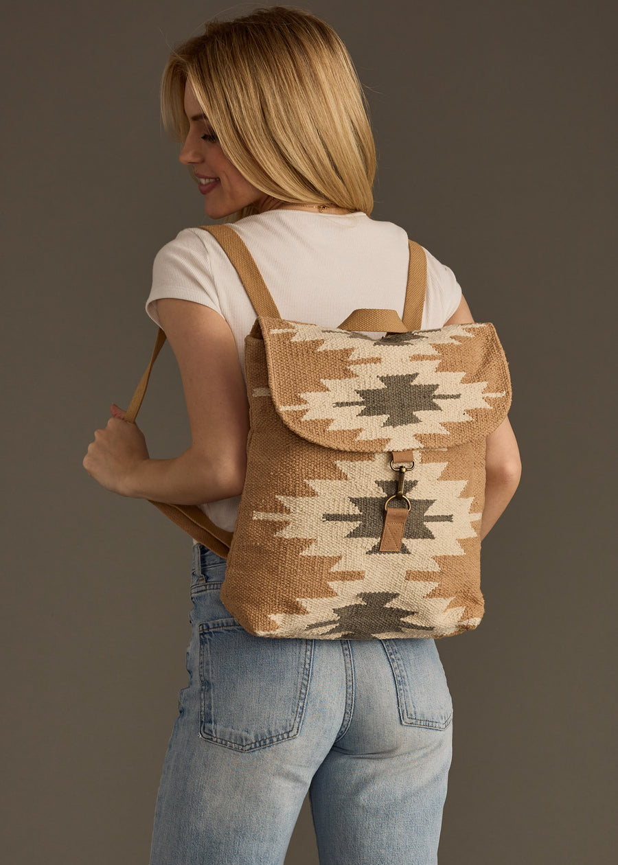 A woman in a white tee and light blue jeans smiles sideways with the Zola Backpack by panacheaccessories—featuring a tan and cream western pattern and adjustable straps—on her back against a plain background.
