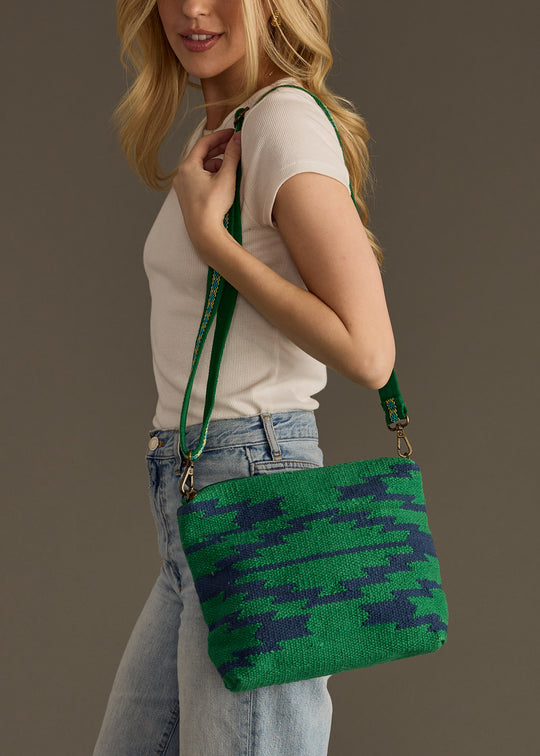 A woman with long blonde hair wears a white short-sleeve top and light blue jeans, carrying the Tilly Crossbody by panacheaccessories in green and navy houndstooth with an adjustable strap. The background is plain gray.