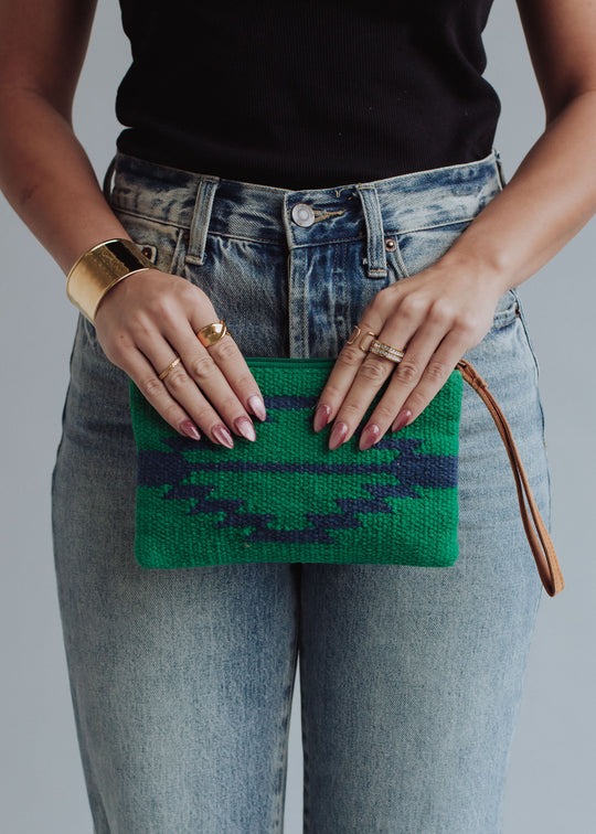 A person in a black top and high-waisted jeans holds the panacheaccessories Tilly Wristlet, a small green and blue patterned clutch with a leather pull. They accessorize with gold rings, a wide gold cuff, and have neatly manicured nails.