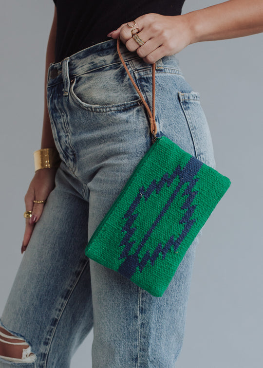 Wearing high-waisted, light-wash jeans and a black top, the person holds the panacheaccessories Tilly Wristlet—a green woven clutch with blue geometric pattern, cotton lining, and tan wrist strap—plus rings and a gold cuff bracelet.