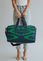 A woman in light blue ripped jeans and nude heels carries the Ebony Duffel by panacheaccessories, featuring green geometric patterns, zipper closure, two handles, a shoulder strap, and soft cotton lining. Plain light gray background.