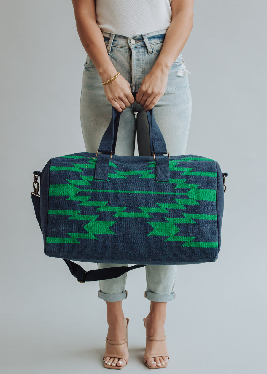 A woman in light blue ripped jeans and nude heels carries the Ebony Duffel by panacheaccessories, featuring green geometric patterns, zipper closure, two handles, a shoulder strap, and soft cotton lining. Plain light gray background.