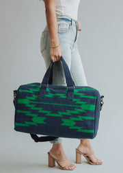 A person in light blue jeans and nude heels carries the panacheaccessories Ebony Duffel, featuring a cotton lining, large navy blue and green pattern, short handles, and a detachable shoulder strap against a plain gray background.