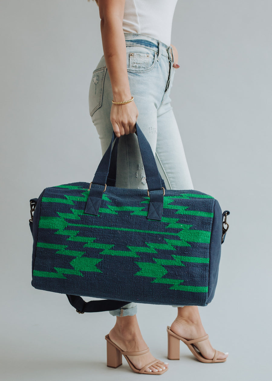A person in light blue jeans and nude heels carries the panacheaccessories Ebony Duffel, featuring a cotton lining, large navy blue and green pattern, short handles, and a detachable shoulder strap against a plain gray background.