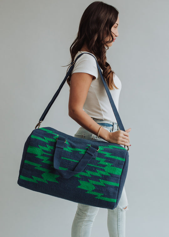 A woman in a white t-shirt and light jeans stands sideways holding the panacheaccessories Ebony Duffel—a large blue and green patterned western bag with shoulder and hand straps, plus a secure zipper—against a plain light background.