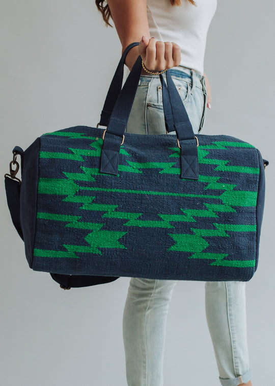 A person in a white sleeveless top and light blue jeans holds the Ebony Duffel by panacheaccessories, featuring navy-blue color, green zigzag patterns, black straps, and a secure zipper closure. The background is plain and light.