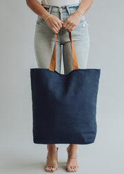 A person in light blue jeans and beige heeled sandals holds the Ebony Tote by panacheaccessories—a large navy bag with brown handles and a playful navy pom tassel—in front of their legs against a neutral background.