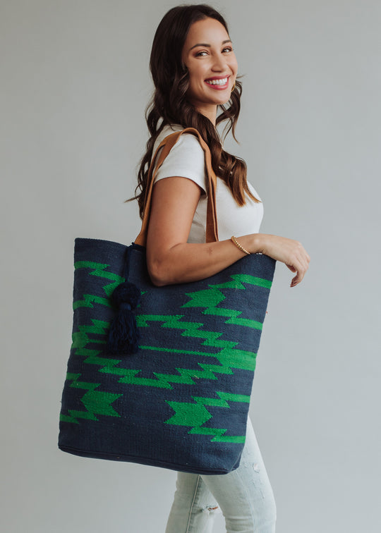 A woman with long brown hair, in a white t-shirt and light jeans, smiles while carrying the Ebony Tote by panacheaccessories—featuring tan leather suede straps and a blue-green patterned design—over her shoulder against a plain background.