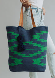 A person in light blue jeans and a white top holds the Ebony Tote by panacheaccessories, featuring green geometric patterns, brown leather handles, and a navy pom tassel, against a plain background.