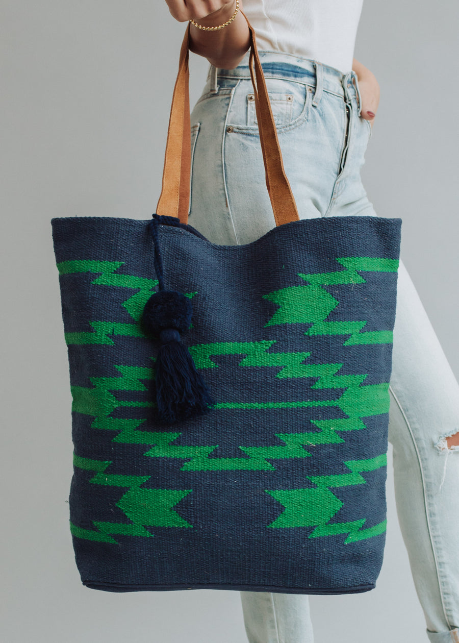 A person in light blue jeans and a white top holds the Ebony Tote by panacheaccessories, featuring green geometric patterns, brown leather handles, and a navy pom tassel, against a plain background.