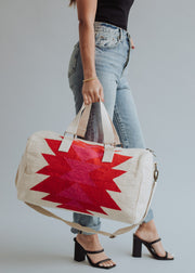 A person in jeans, a black top, and heeled sandals holds the panacheaccessories Stella Duffel—a cream-colored, western-inspired bag with red geometric patterns, cotton lining, handles, shoulder strap, gold hardware, and zipper closure. Plain gray background.